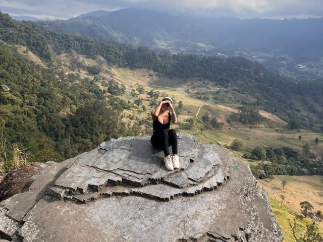 MajoJones seduta su una grande roccia grigia, vestita di nero e con scarpe da ginnastica bianche, con i seni coperti dalle mani, che guarda la fotocamera, in un paesaggio montano..