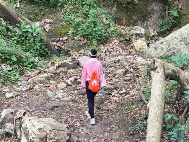 MajoJones, portant une chemise rose, un pantalon noir et des baskets blanches, et portant un sac à dos rouge, marchant sur un sentier forestier rocheux et ombragé, porte un chapeau noir..