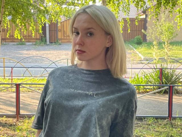 Carmela Bonuz mit blonden Haaren, die ein graues T-Shirt im Batik-Stil trägt und einen ernsten Gesichtsausdruck hat, steht im Freien vor einem Metallzaun und viel Grün im Hintergrund. Ihre Brüste sind klein, und sie ist niedlich..