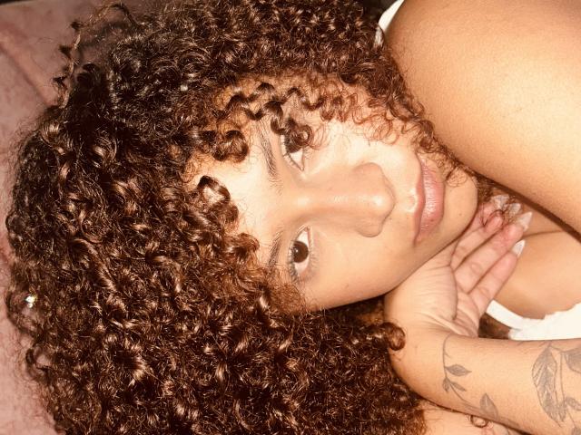 Sofia a les cheveux bruns bouclés, une peau brun clair, et elle est allongée, la tête reposant sur sa main. Ses tatouages sont visibles sur son bras droit..