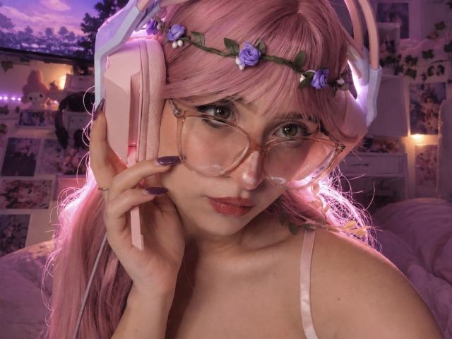 ViviCute, con cabello rosa, una corona de flores, auriculares rosas, gafas grandes y transparentes, y una camiseta rosa con tirantes, sosteniendo su auricular, mostrando su cara adorable y su lápiz labial rosa..