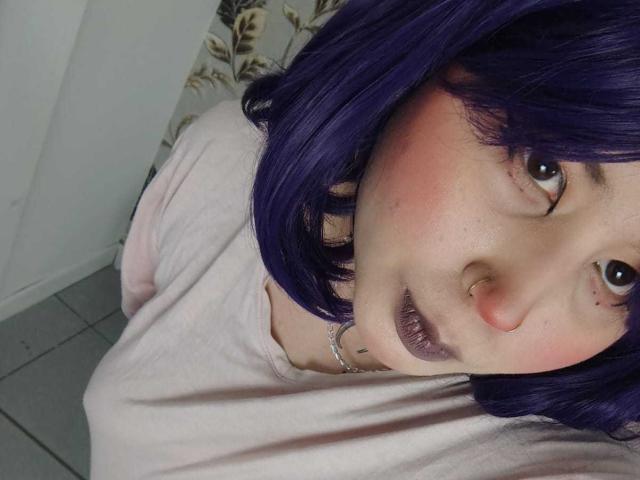 WendyMonet with purple hair, pink blush, nose ring, and purple lipstick, wearing a pink shirt, looking up at the camera, showing a cute, slightly mischievous face.