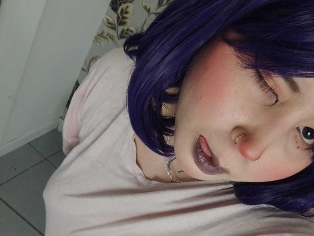 WendyMonet with purple hair, pink cheeks, nose ring, and dark lipstick, wearing a pink shirt, looking up with one eye closed, showing a subtle lip piercing.