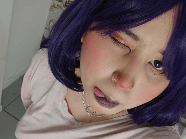 WendyMonet with purple hair, winking, tongue out, pink nose, silver chain necklace, and purple lipstick, wearing a pink shirt, on a gray floor.