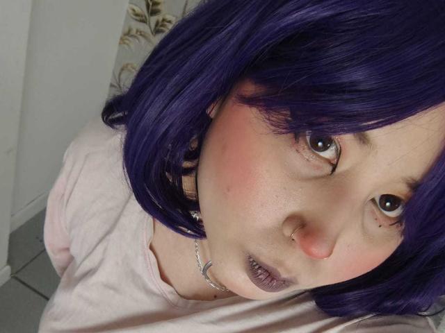 WendyMonet with purple hair, pink blush, and purple lipstick, wearing a pink shirt, has a nose ring and a silver chain necklace, looking up with wide eyes.