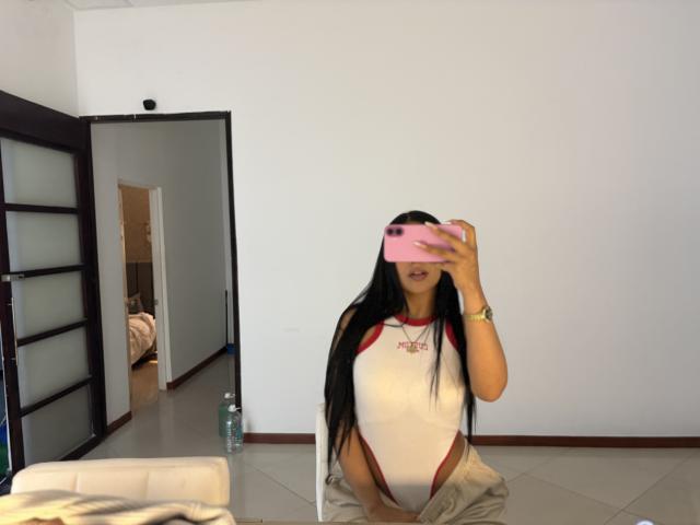 Antonella Monroe, avec de longs cheveux noirs, portant une chemise raglan blanche et rouge, un pantalon beige, tenant un téléphone rose, autoportrait pris dans un miroir, visage et seins visibles, pièce avec une porte ouverte..