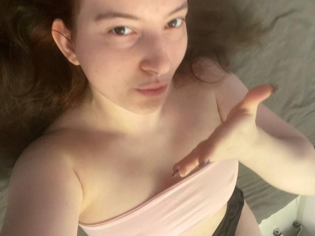 JohnnaChelf ligt op een bed, gekleed in een roze top zonder bandjes, waarbij haar kleine borsten zichtbaar zijn. Ze heeft bruin haar, een neusring en een hand die omhoog wijst, terwijl ze naar de camera kijkt..