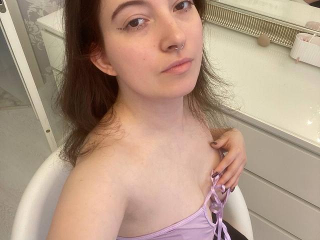 JohnnaChelf, con la pelle chiara, i capelli castani e un piercing al naso, tiene una tracolla viola, mostrando una spalla scoperta e piccoli seni, seduta su una sedia bianca, mentre guarda la fotocamera..