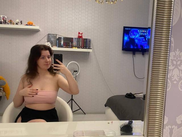 JohnnaChelf, avec des seins couverts par un haut rose sans bretelles, une jupe noire, tenant un téléphone dans un selfie pris devant un miroir, une chaise blanche, des cheveux bruns, un anneau lumineux, une étagère murale, une télévision affichant un écran bleu..