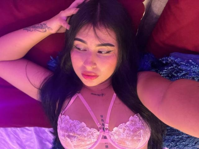 Hannah Becker, met lang zwart haar, een roze kant beha en tatoeages, liggend op rode kussens, met haar hand in haar haar, naar beneden kijkend, met roze en paarse verlichting, een zachte uitdrukking, volle, roze lippen..
