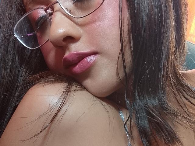 SweetDeya's face with glossy pink lips, small nose piercing, and round glasses, showing a slight pout. She has long brown hair and a shiny, smooth complexion.