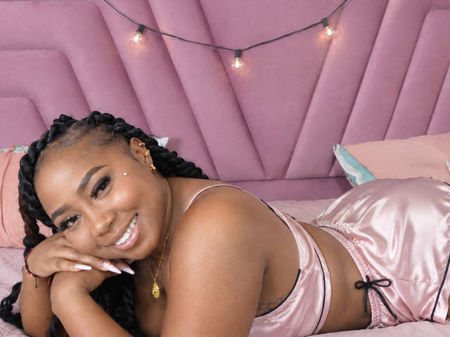 DarkSkinnedTaboo mit glänzendem, rosa Seidendessous, liegt auf einem rosa Bett, lächelt, hat geflochtene Haare, goldene Ohrringe, und darüber hängen Lichterketten, wobei auf dem Foto ihre Brüste und ihr Hintern zu sehen sind..
