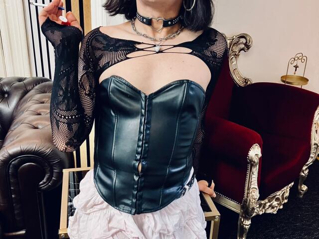 FemCupid in a black lace top, green leather corset, and white ruffled skirt, holding a cigarette, with a black choker and silver ring. Her tits are small, barely visible.
