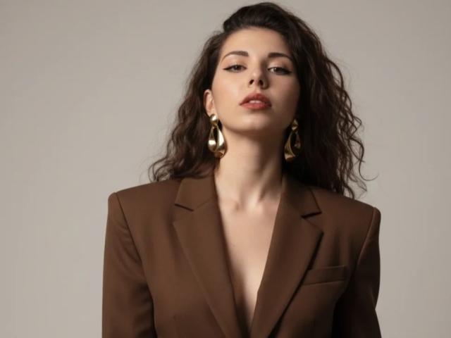 DellaSwartzbaugh with wavy dark hair, brown blazer, deep V-neck, gold earrings, looking slightly up, soft expression, slightly parted red lips. Boobs not visible, focus on upper body.