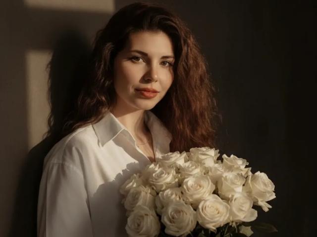 DellaSwartzbaugh with curly brown hair, white shirt, holding a bouquet of white roses, soft light, slight smile, looking at camera, subtle shadow on wall.