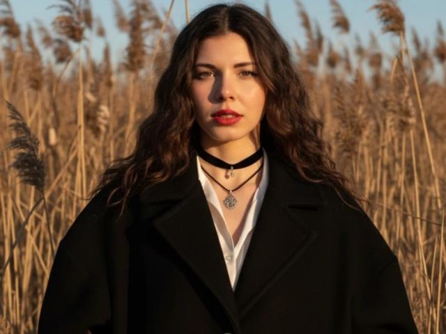 DellaSwartzbaugh with wavy brown hair, red lipstick, black choker, black coat, white shirt, and pendant, standing in a tall grass field, looking serious, in warm sunlight.