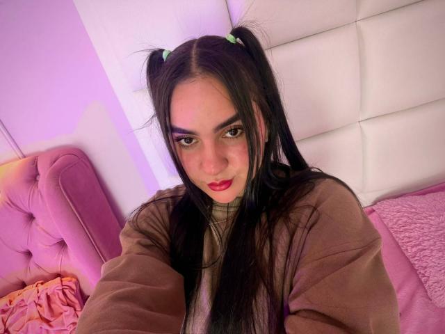 FenixAlthea with long black pigtails, pink eyeshadow, red lipstick, brown sweater, and cute expression, sitting on pink bed with tufted headboard, looking at camera.