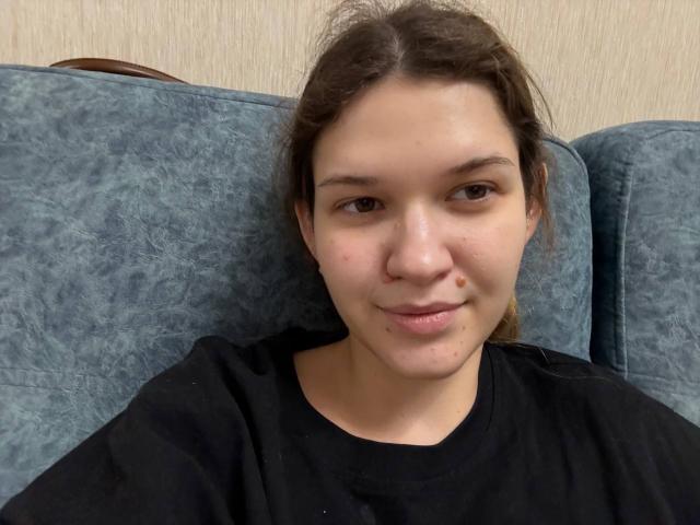Georgan Hildman, mit heller Haut, braunen Augen und einer kleinen Pickel auf der rechten Wange, trägt ein schwarzes Hemd und sitzt auf einem blau gemusterten Sofa, wobei er leicht nach links schaut..