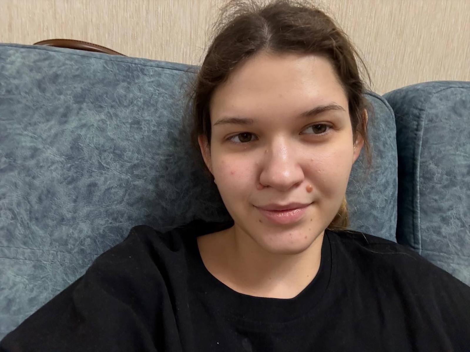 Georgan Hildman, mit heller Haut, braunen Augen und einer kleinen Pickel auf der rechten Wange, trägt ein schwarzes Hemd und sitzt auf einem blau gemusterten Sofa, wobei er leicht nach links schaut..