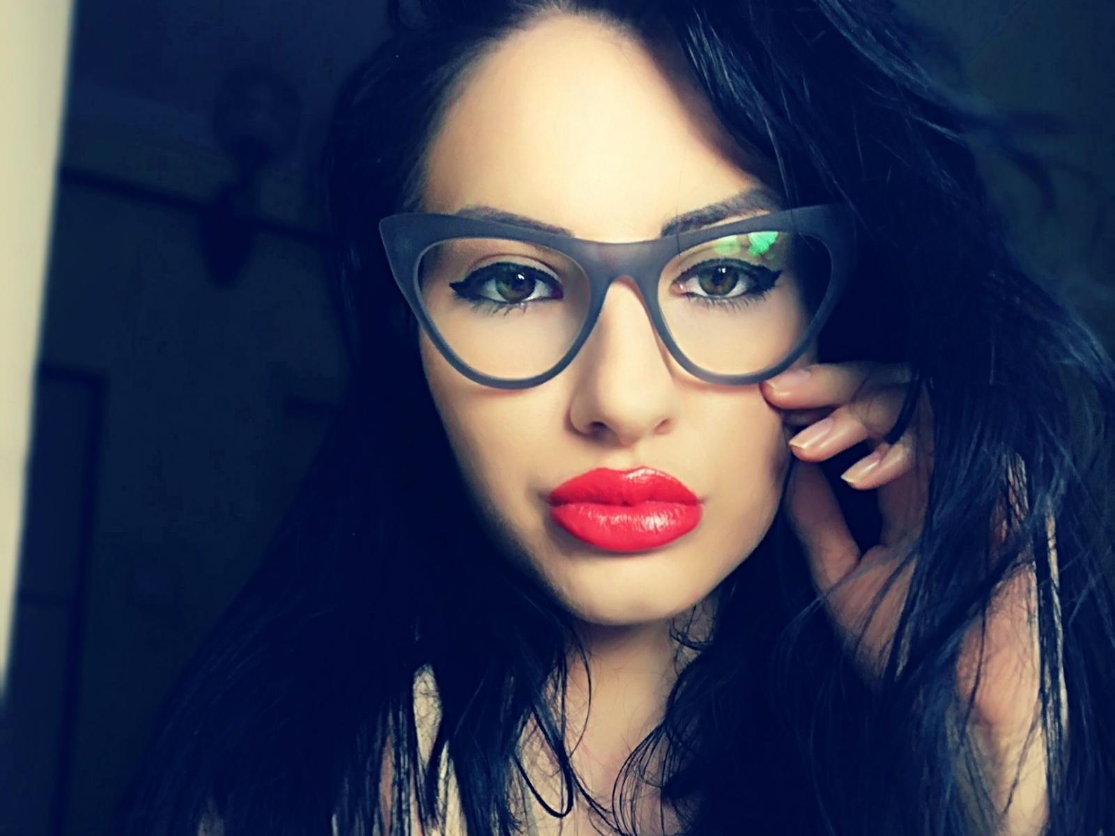 Zaccada with red lipstick, green eyes, and black hair, wearing big gray glasses, touching her cheek with her hand, looking into the camera. Her tits are not visible.