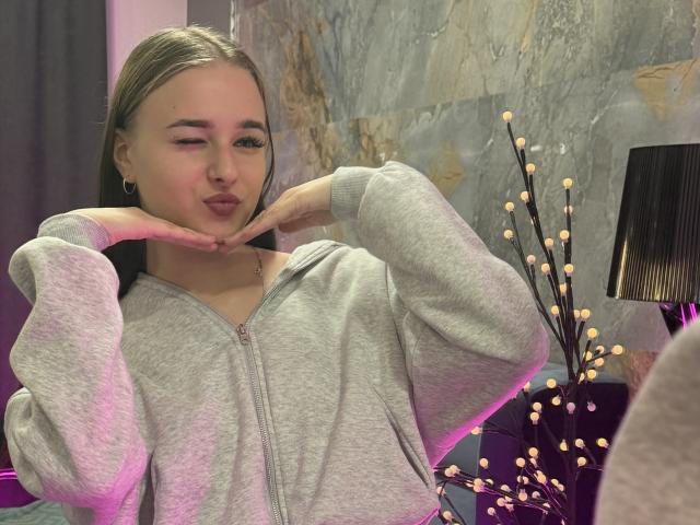 Lixsi with pouty lips, hands under chin, wearing a grey zip-up hoodie, blonde hair, slight pink lighting, black lamp, and decorative branches with lights in the background.