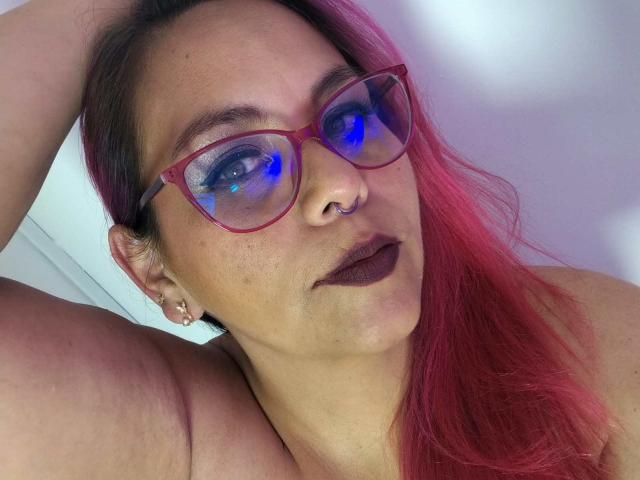 CamilleFoxyHot con i capelli rosa, occhiali rossi che riflettono luce blu, piercing al naso, rossetto scuro e orecchini, in posa con un braccio dietro la testa, mostrando una spalla scoperta..