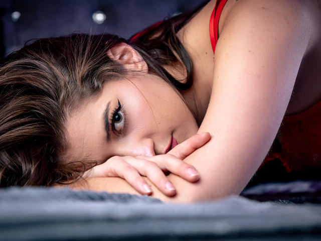 Lana Sanders ligt op haar zij met haar gezicht rustend op haar arm, ze draagt een rode top en kijkt met een subtiele glimlach naar de camera. Haar borsten zijn nauwelijks zichtbaar..