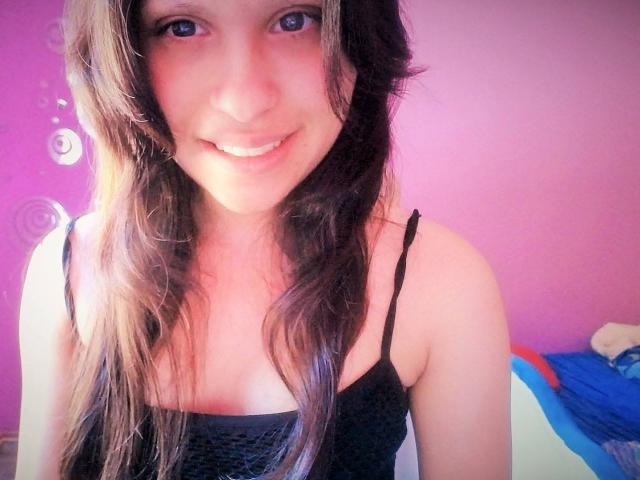 AngelinaHottiestX, con cabello castaño largo, ojos azules y un top negro con tirantes finos, sonriendo a la cámara, mostrando sus lindos senos y un ligero bronceado en su piel clara..