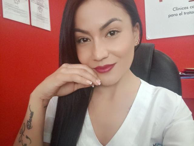 NinnaBallack with long black hair, light skin, and pink lipstick, wearing a white V-neck shirt, has a tattoo on her right wrist and is smiling with her hand near her mouth.