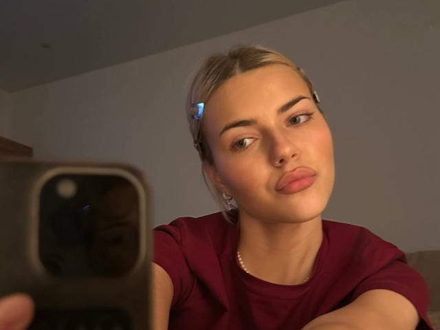 ArnettaNazario with blonde hair in a clip, pouty lips, and a nose ring, wearing a maroon shirt, taking a selfie with a phone in a dimly lit room.