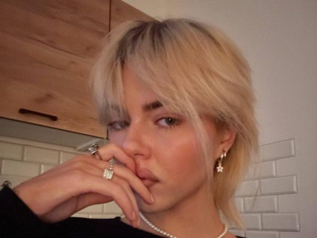 Arnetta Nazario, avec ses cheveux blonds, ses lèvres pulpeuses, portant un haut noir, un collier de perles et des boucles d'oreilles en forme de croix, touchant son nez avec une main ornée de bagues, avec un regard séduisant..