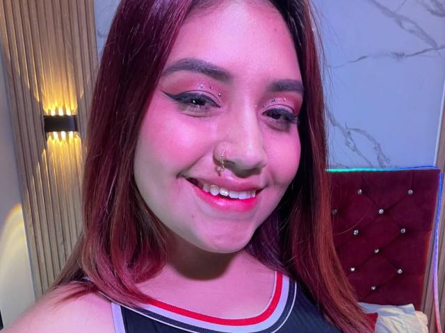 ValmontAlessia with red hair, nose piercing, and eyeliner, smiling at the camera. She's wearing a black and red top, with pink lipstick and a cute face.