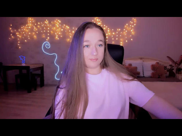 NikkaVeira con lunghi capelli castani, una camicia rosa, seduta su una sedia nera, una linea al neon blu sulla parete, luci decorative calde, pavimento in legno, un piccolo tavolo e un orsacchiotto sullo sfondo..