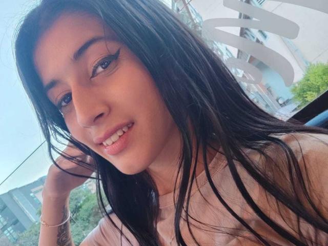 JasminKooper, con cabello negro largo, vestida con una blusa rosa, tiene delineador de ojos alado, está sonriendo y tiene una mano en el cabello, luciendo linda y sexy..