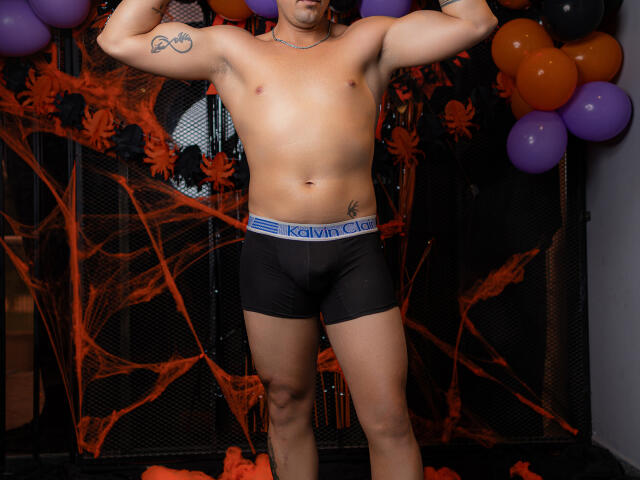 ElkynAndNell, shirtless, flexing arms, wearing black Calvin Klein briefs, tattoo on left bicep, purple waistband, standing in front of Halloween decorations, balloons in background.