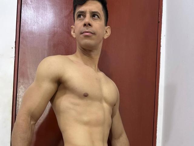 JoseRumBerotop, sem camisa, com um peito musculoso e definido, e abdominais visíveis, em frente a uma porta vermelha, olhando ligeiramente para o lado com uma expressão neutra..