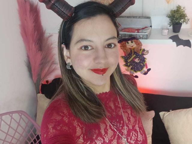 ArianaBlonde with brown hair, red lace top, and devil horns, smiling, red lipstick, small earrings, necklace, pink wall, and pink decorative feather in the background.