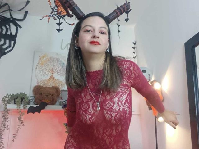 ArianaBlonde with straight brown hair, wearing a red lace dress, red lipstick, and earrings, standing with arms slightly out, looking at the camera, in a Halloween-themed room.
