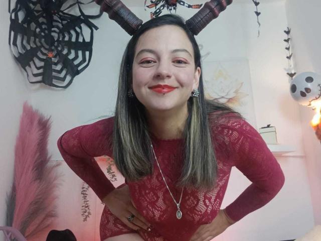 ArianaBlonde with black horns, red lace bodysuit, and red lipstick, smiling with hands on hips, wearing a necklace with a circular pendant, and striped earrings.