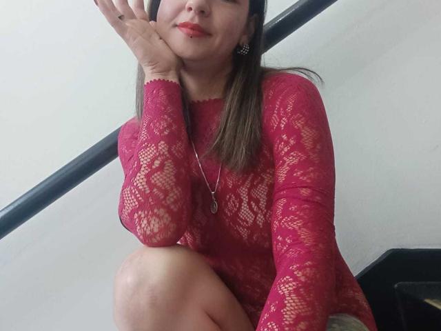 ArianaBlonde in a pink lace top, squatting with one knee up, hand on cheek, red lipstick, brown hair, silver necklace, black ear studs, smiling subtly.