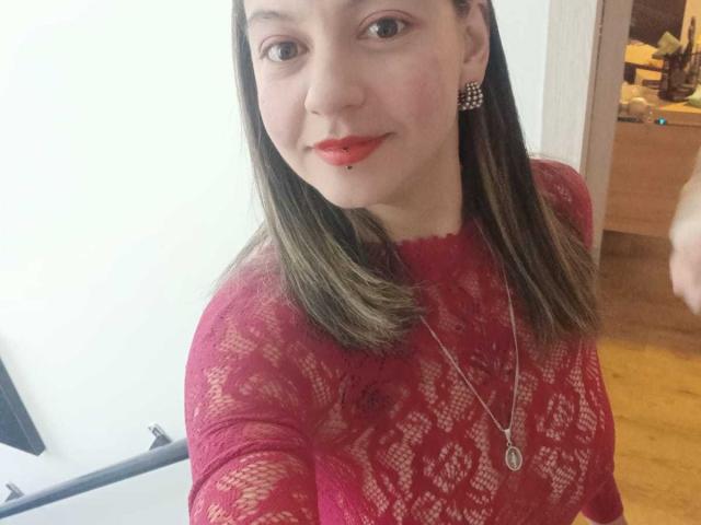 ArianaBlonde with red lace top, straight brown hair, red lipstick, nose piercing, and pearl earrings, smiling at the camera, wearing a silver necklace.