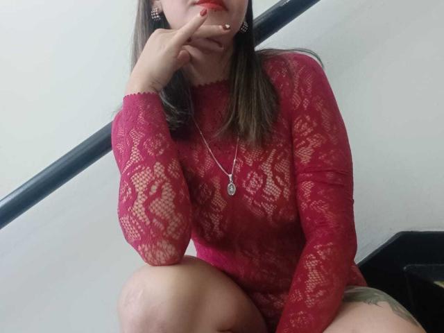 ArianaBlonde with red lace top, showing her tits through the fabric, sitting with knee up, hand on chin, red lipstick, necklace, and tattoo on thigh.
