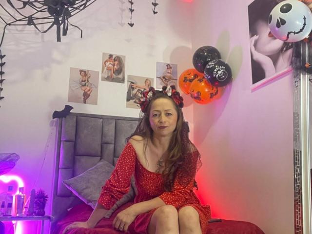 GianineLarose with long brown hair, wearing a red polka dot dress and Mickey ears, sitting on a bed with a gray headboard, surrounded by Halloween decorations and balloons.