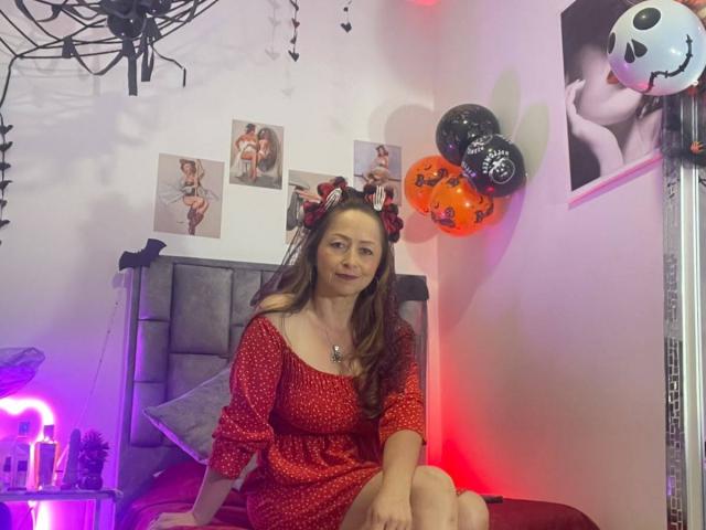 GianineLarose, with long brown hair, red polka-dot dress, and Halloween headband, sits on bed, legs crossed, in room with purple and red lights, Halloween balloons, and sexy posters.