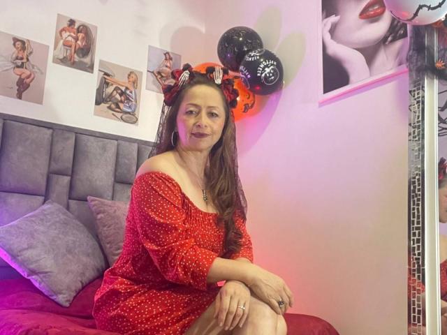 GianineLarose, with long brown hair and red polka-dot dress, sits on a bed wearing Mickey Mouse ears, her ass barely covered, smiling in a pink-lit room.