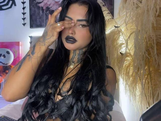 LairaHearvy with long black hair, tattoos on her hand and neck, dark lipstick, covering one eye, wearing a black lace top, in a bedroom with puffy gold curtains.