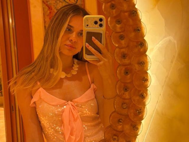 Sammie Scantlen mit langen blonden Haaren, einem pinkfarbenen Top mit Pailletten und einer weißen Muschelkette, macht ein Selfie im Spiegel und zeigt ihre Brüste. Sie hält ein goldenes iPhone in einem warm beleuchteten Raum..