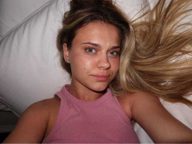 SammieScantlen lying on a white pillow, wearing a pink sleeveless top, with long blonde hair spread out, green eyes, and a small beauty mark on her right cheek.