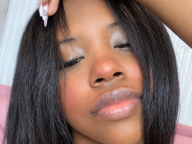 Le visage de CatalinaSanA, les yeux fermés, avec des lèvres roses brillantes, des cheveux noirs et lisses, et une main touchant sa tête, mettant en valeur son ongle rose avec un bijou..