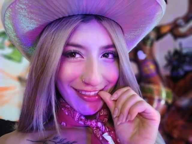 Kaiix, com cabelos loiros longos, um chapéu roxo brilhante, um piercing rosa no nariz e um lenço vermelho na cabeça, sorrindo e mordendo o dedo. Seus seios estão parcialmente visíveis..